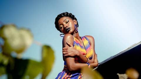 A Black woman wearing a colorful dress and hoop earrings with her eyes closed, hugging herself