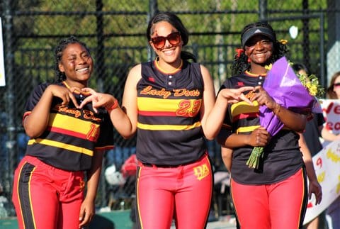 Three smiling softball players, all young Black women. One is making a shape of a heart with her hands, one is wearing sunglasses, and one is holding a bouquet of flowers.
