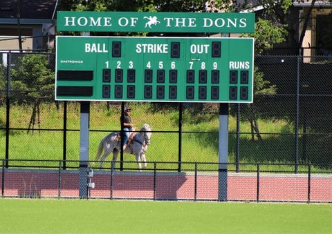 A scoreboard that says Home of the Dons. Beyond the scoreboard and the chain-link fence behind it is a person on a horse.