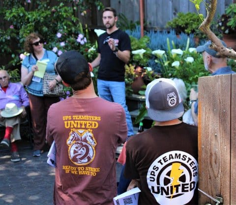 Two men wearing T-shirts that say "UPS Teamsters United" on the back watch another man speak at an outdoor event. One man's shirt also says "ready to strike if we have to."