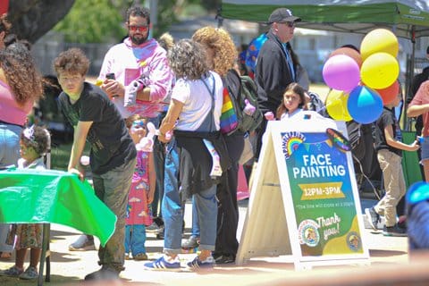 A sign that says "Pinole Pride" and "face painting" with balloons tied to it among a crowd
