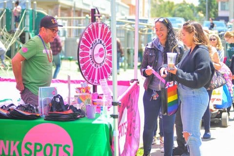 Two women stand at a booth staffed by a man. The booth has a pink and green color scheme and a prize wheel