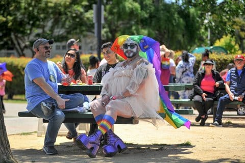 A drag queen wearing a rainbow headscarf, white face makeup, sunglasses, dress, jewelry and platform boots sits at a park bench with other people.