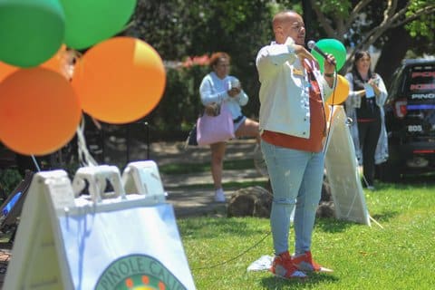 A younger Black man speaking with a microphone in a park. Green and orange balloons are tied to a sandwich board sign nearby