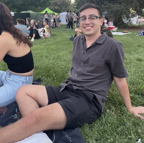 A smiling young Latino man wearing glasses and sitting on grass among a crowd