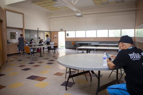 A man in a black T-shirt that says Whole Foods Market on it, a black ball cap and a blue surgical mask sits at a table with a bottle of water looking at his phone. A few other people are at another table across the room.