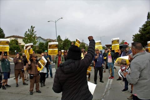 A Black person seen from behind raises their fist while facing a group of people, many holding picket signs that read "just practicing for a just contract"