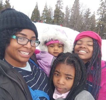 A smiling Black woman with her three daughters (aged 2, 10 and 13) in a snowy area with tall trees in the background