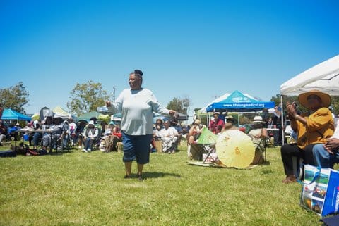 A Black woman dances on grass during a festival. There are several other people sitting around.