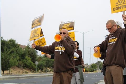 A bald white man using a cone-shaped device to amplify his voice stands roadside with a sign that says "UPS teamsters just practicing for a just contract." He is wearing a brown sweatshirt that says "ready to strike if we have to." With him are other people with the same signs and sweatshirts.