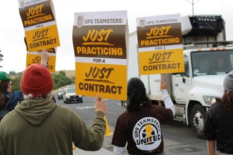 People seen from behind next to a street while holding picket signs that read "just practicing for a just contract." One person's shirt says "UPS teamsters united" on the back