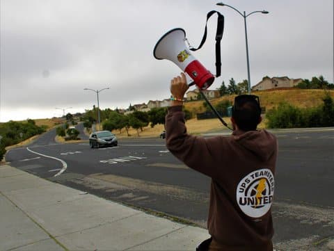 A man seen from behind stands roadside holding a megaphone aloft. He is wearing a brown sweatshirt that says "UPS teamsters united" on the back. One car is on the road.
