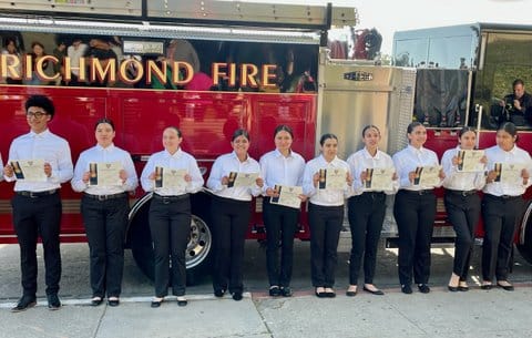 Ten teens dressed in white button-down shirts and black pants holding up certificates in front of a Richmond Fire truck