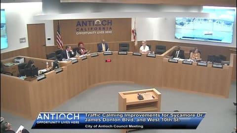 Antioch City Council meeting with six people sitting in elevated seats in front of a sign that says Antioch California opportunity lives here. On screen text reads traffic improvements for sycamore drive, james donlon boulevard and west 10th street