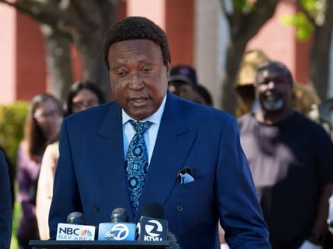 Attorney John Burris, a Black man in a suit, stands in front of microphones for NBC Bay Area, ABC 7 and KTVU 2