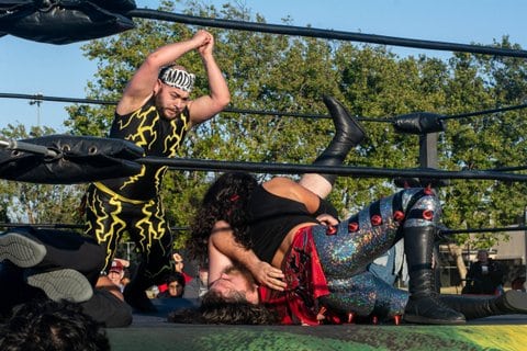 In the ring, one wrestler wearing a black unitard with yellow lightning bolt designs holds a fist clasped in his other hand above two other fighters. One is wearing silver-sequined pants with red spikes and tall, black boots.