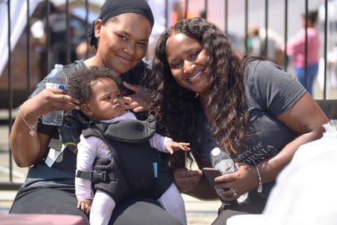 Two smiling Black women. One is wearing a do-rag and has a Black baby strapped to her front. The other has long, curly hair.