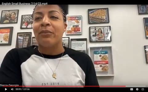 Screenshot of a Black woman sitting in front of several framed pictures and articles