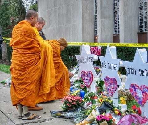 Three Buddhist monks, dressed in orange robes, at a memorial for victims of the Tree of Life synagogue shooting. There are two markers shaped like the Star of David with the names Richard Gottfried and Rose Mallinger and a third that appears to show the name Jerry Rabinowitz, along with flowers, heart decals and candles.
