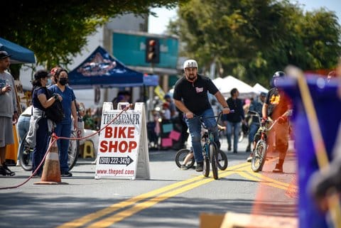 A man rides his bike during a street festival past a sign that reads The Pedaler El Sobrante bike shop. 510 222 3420. w w w dot the ped dot com