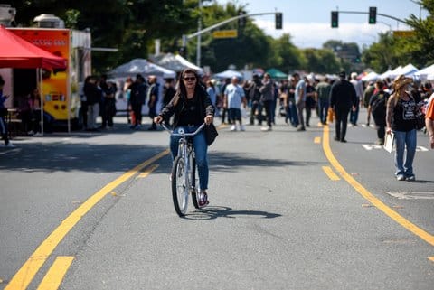 A woman rides a bike on a street closed for an event. Many people are walking on the street and some stands are visible.