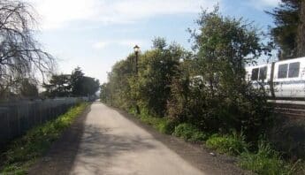 A paved path lined with greenery, including trees on one side, and a Bart train running alongside.
