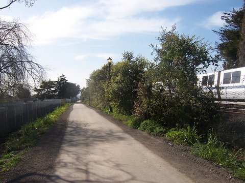 A paved path lined with greenery, including trees on one side, and a Bart train running alongside.