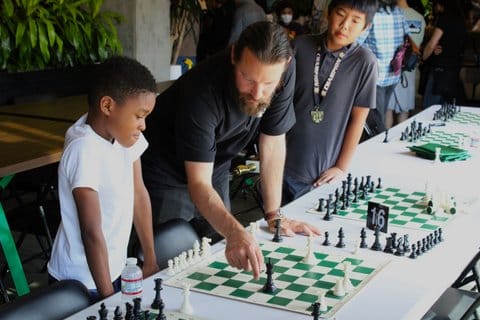 A man gestures toward a chess board while a boy looks on