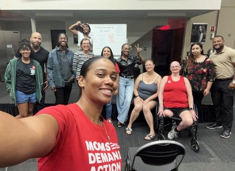A smiling Black woman in a red T-shirt that says "moms demand action" takes a selfie with a group of people behind her
