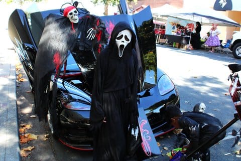 A kid in a Ghostface costume stands in front of a black car decorated for Halloween