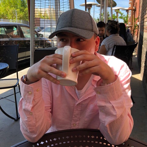 A Latino young man in a pink shirt sipping from a coffee mug while sitting outside