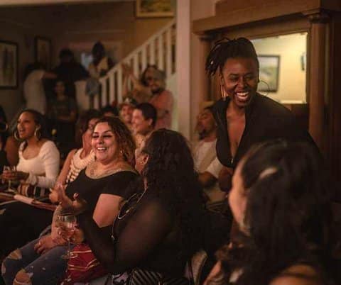 A group of people in the audience in a house-like setting. Shown most prominently are a Latina woman and a Black woman, both with joyful expressions on their faces
