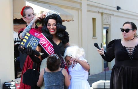 A woman in a black dress with black gloves, styled black hair, tiara and a film clapper. She is wearing a sash for winning a pin-up girl costume contest. Two little girls and two adults in dresses are standing around her.
