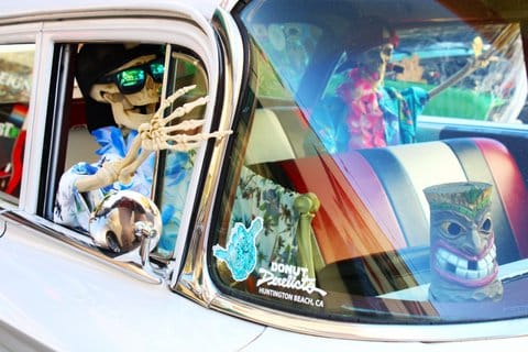A car with skeletons in the passenger and back seats. They are wearing Hawaiian shirts, leis and backwards caps. One has sunglasses. There is a tiki mug on the dashboard