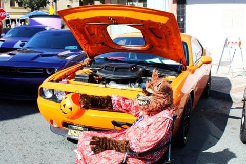 A person dressed up as the big bad wolf as grandma sitting in front of an orange muscle car holding out a plastic jack-o-lantern bucket