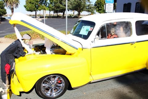 A yellow car with its hood up and fake figures looking under the hood and sitting in the driver's seat