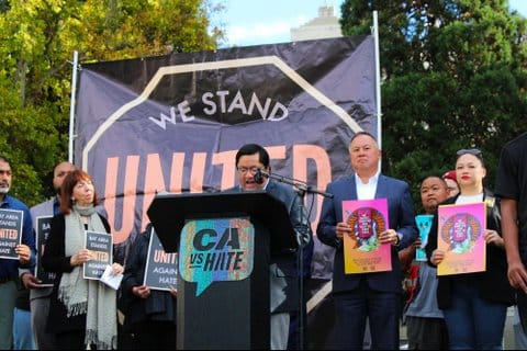A Latino man speaking at a lectern with a sign that says CA VS Hate. People on stage with him hold posters that say "bay area stands united against hate" or share another anti-hate message in a non-English language. On a backdrop behind them, the words "we stand united" are visible