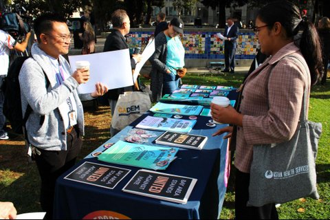 An Asian man smiles at a South Asian woman across a table in a park as she looks down at posters laid out that say "bay area stands united against hate" and other anti-hate messages. Her bag says "asian health services." Other people are in the background.