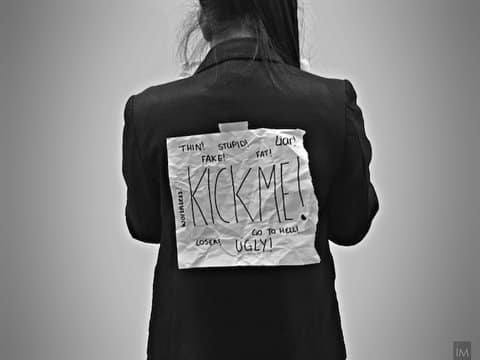 Person with paper taped to their back that says "kick me" in large lettering. Other writing on the paper says thin, stupid, liar, fat, fake, worthless, ugly, go to hell