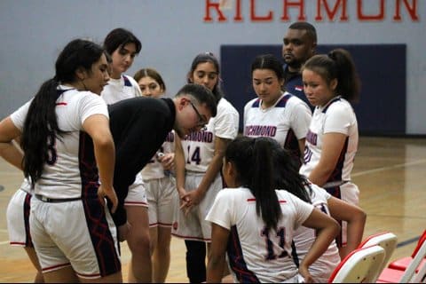 Girls basketball players and a male coach standing around the bench area with two players seated each with a hand on their backs.