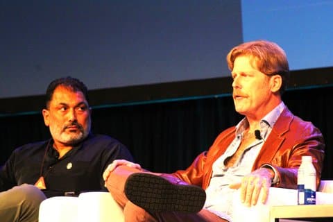 An Indian American man with dark hair and mostly gray beard looking at a white man with red hair and mustache. They are seated side by side.
