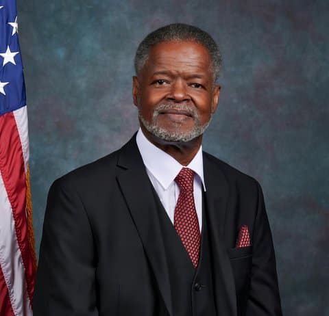 An older, distinguished looking Black man with salt and pepper hair and beard in a suit next to the American flag