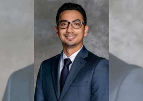 A smiling young Latino man in a suit and glasses