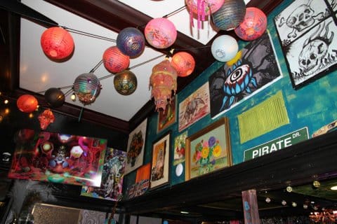Looking up toward a ceiling decorated with paper lanterns in different colors. Also on the walls are artworks depicting flowers, skulls, a street sign for Pirate Drive and more.
