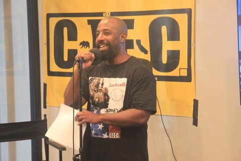 A smiling Black man with a microphone who is wearing a T-shirt that has the word Juneteenth and a U.S. flag as part of its design