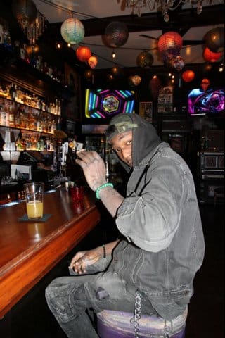 A young Black man sitting at a bar looking sideways toward the camera with one hand raised. He is wearing a backwards cap and a hoodie. The hood is slightly covering his face.