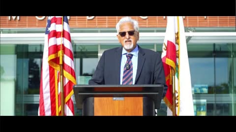 A gray-haired Latino man in sunglasses and suit and tie standing at a lectern between the U.S. and California flags