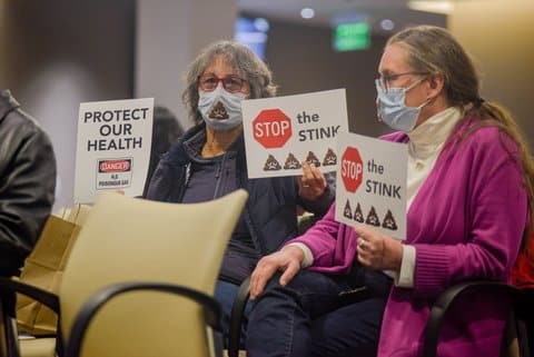 Two older white women at a city council meeting wearing glasses and surgical masks decorated with a poop emoji. One is looking at the camera and holding up signs that read "protect our health. danger, poisonous gas" and "stop the stink" with a stop sign and poop emojis. The other woman is looking away and holding a stop the stink/poop sign