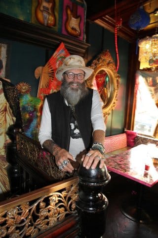 A smiling white man with a long, gray beard. He is wearing a hat, glasses and several rings and bracelets and has tattoos on his arms.