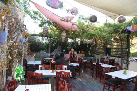 A restaurant patio with several tables. A white woman is standing by one where a Black person is seated. Large pieces of cloth, paper lanterns and upside-down parasols are strung up above.